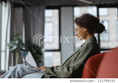 A woman comfortably sitting on a couch with a laptop computer in nice modern coworking office A woman comfortably sitting on a couch with a laptop computer in nice modern coworking office 112799141