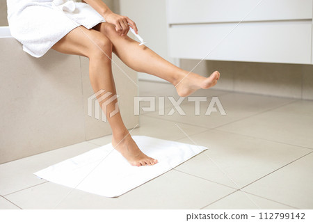 A woman sits by a window shaving her leg on hardwood flooring 112799142