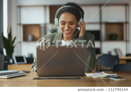 A woman with headphones smiles at her laptop on the table in nice modern coworking office A woman with headphones smiles at her laptop on the table in nice modern coworking office 112799144