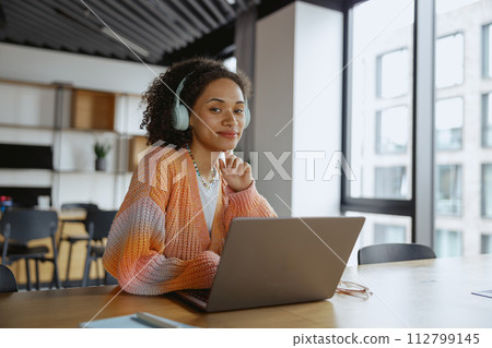 Woman with headphones working on laptop at table in a building with windows Woman with headphones working on laptop at table in a building with windows 112799145