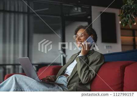 a business woman is sitting on a couch using a laptop and talking on a cell phone in nice modern coworking office 112799157