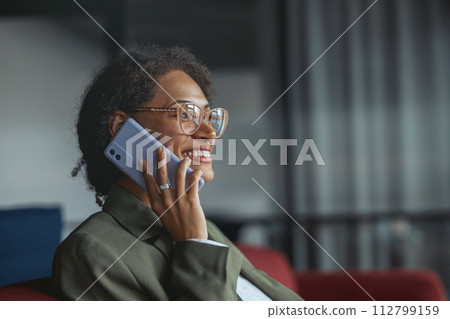 A business woman with glasses sitting on a couch talking on a cell phone in nice modern coworking office A business woman with glasses sitting on a couch talking on a cell phone in nice modern coworking office 112799159