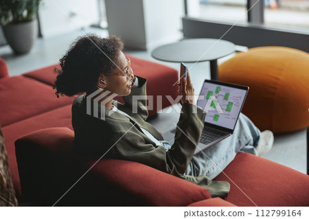 Woman lounging comfortably on red couch, typing on laptop with fingers in nice modern coworking office Woman lounging comfortably on red couch, typing on laptop with fingers in nice modern coworking office 112799164