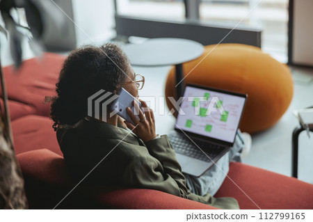 Woman on the couch with a laptop, talking on phone in nice modern coworking office Woman on the couch with a laptop, talking on phone in nice modern coworking office 112799165