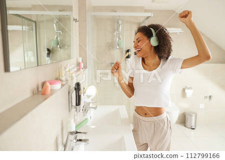 A woman with headphones brushes her teeth in the bathroom, smiling happily 112799166