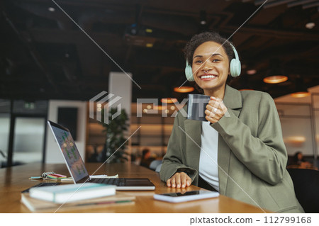 A woman at a desk wearing headphones smiles while drinking coffee and working in nice modern coworking office 112799168