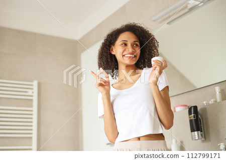 A woman gazes at her reflection in the mirror, standing in the bathroom A woman gazes at her reflection in the mirror, standing in the bathroom 112799171