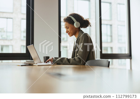 A woman with headphones sits at a table using a laptop computer in nice modern coworking office A woman with headphones sits at a table using a laptop computer in nice modern coworking office 112799173