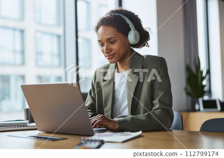 A woman with headphones is at a table using a laptop computer A woman with headphones is at a table using a laptop computer 112799174
