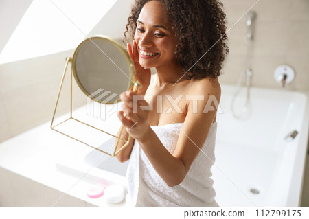 A woman with a towel around her body admires herself in the bathroom mirror 112799175