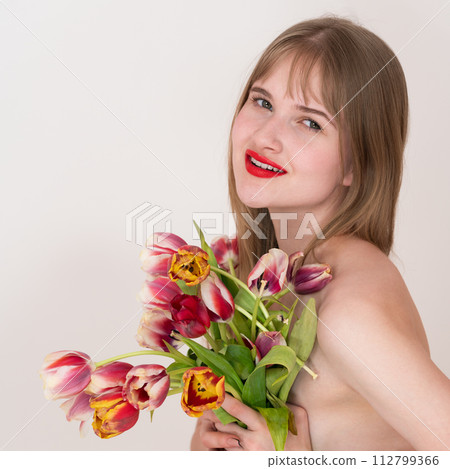 Portrait of blonde woman holding stunning bouquet of spring tulips and playfully covering her chest with them while smiling and gazing right at camera. Lovely young blonde woman with bright red lips Portrait of blonde woman holding stunning bouquet of spring tulips and playfully covering her chest with them while smiling and gazing right at camera. Lovely young blonde woman with bright red lips 112799366