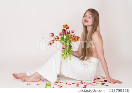 Half naked woman holding bouquet of spring tulips flowers for Woman's Day with closed eyes. Serene blonde woman with long hair falling down and covering her chest. Model sitting on white background Half naked woman holding bouquet of spring tulips flowers for Woman's Day with closed eyes. Serene blonde woman with long hair falling down and covering her chest. Model sitting on white background 112799368