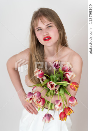 Portrait of blonde woman holding bouquet of tulips, using them to cover her chest in playful and flirty way. Her long hair cascading down chest, she looking at camera with confident and alluring gaze Portrait of blonde woman holding bouquet of tulips, using them to cover her chest in playful and flirty way. Her long hair cascading down chest, she looking at camera with confident and alluring gaze 112799369