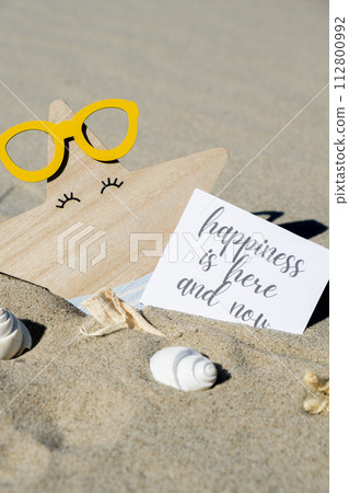 HAPPINESS IS HERE AND NOW text on paper greeting card on background of funny starfish in glasses summer vacation decor. Sandy beach sun coast. Slowing-down, enjoying the moment, good moments, slow 112800992