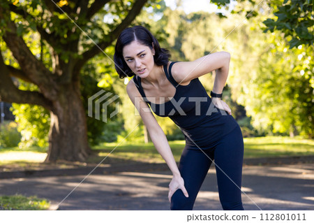 Upset young woman standing in the park bent over and holding her hand behind her back, suffering from severe pain and sprain after jogging and working out. Upset young woman standing in the park bent over and holding her hand behind her back, suffering from severe pain and sprain after jogging and working out. 112801011