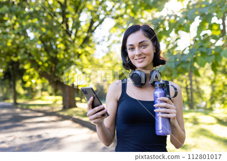 Portrait of a young female athlete and trainer standing in the park wearing headphones, holding a phone and a bottle of water in her hands, looking at the camera with a smile. 112801017