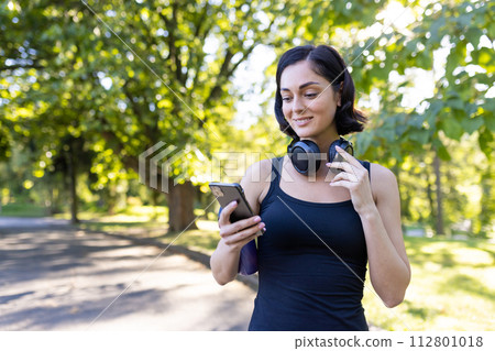Close-up photo of a young girl jogging and doing sports in the park, standing in headphones and using a mobile phone. 112801018