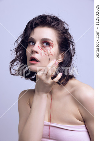 Portrait of beautiful woman with red string on face and hand with needle. Complexities of beauty, facial changes. Portrait of beautiful woman with red string on face and hand with needle. Complexities of beauty, facial changes. 112802028