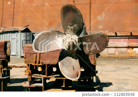 Large, rusted ship propeller displayed on wooden blocks in a shipping yard, symbolizing industrial maritime decommission. Large, rusted ship propeller displayed on wooden blocks in a shipping yard, symbolizing industrial maritime decommission. 112803036
