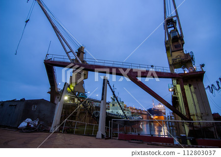 Industrial shipyard at night, floating dock with a ship under repair, lit cranes on duty, marine engineering scene. Ship maintenance, maritime transportation industry, dockyard operations depicted. Industrial shipyard at night, floating dock with a ship under repair, lit cranes on duty, marine engineering scene. Ship maintenance, maritime transportation industry, dockyard operations depicted. 112803037
