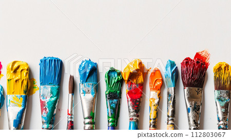 An arrangement of colorful paintbrushes dipped in various hues of paint on a pristine white surface 112803067