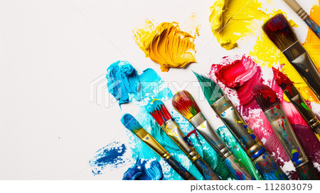 An arrangement of colorful paintbrushes dipped in various hues of paint on a pristine white surface 112803079