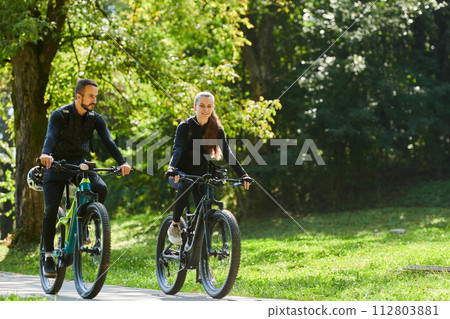 A blissful couple, adorned in professional cycling gear, enjoys a romantic bicycle ride through a park, surrounded by modern natural attractions, radiating love and happiness 112803881