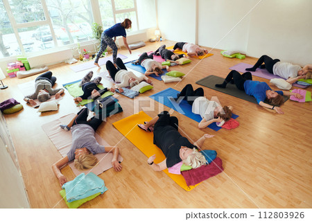 A group of senior women engage in various yoga exercises, including neck, back, and leg stretches, under the guidance of a trainer in a sunlit space, promoting well-being and harmony 112803926