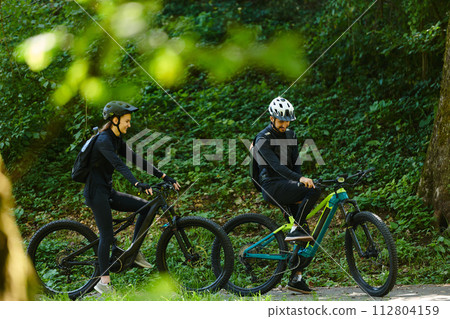 A blissful couple, adorned in professional cycling gear, enjoys a romantic bicycle ride through a park, surrounded by modern natural attractions, radiating love and happiness 112804159