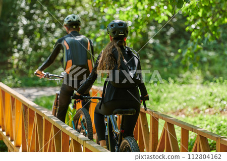 A blissful couple, adorned in professional cycling gear, enjoys a romantic bicycle ride through a park, surrounded by modern natural attractions, radiating love and happiness 112804162