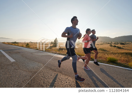 A group of friends, athletes, and joggers embrace the early morning hours as they run through the misty dawn, energized by the rising sun and surrounded by the tranquil beauty of nature A group of friends, athletes, and joggers embrace the early morning hours as they run through the misty dawn, energized by the rising sun and surrounded by the tranquil beauty of nature 112804164
