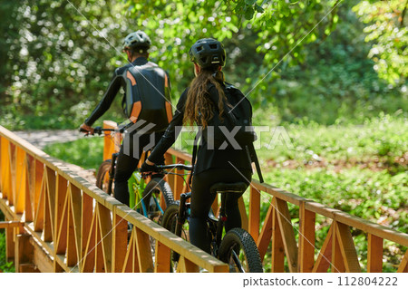 A blissful couple, adorned in professional cycling gear, enjoys a romantic bicycle ride through a park, surrounded by modern natural attractions, radiating love and happiness A blissful couple, adorned in professional cycling gear, enjoys a romantic bicycle ride through a park, surrounded by modern natural attractions, radiating love and happiness 112804222