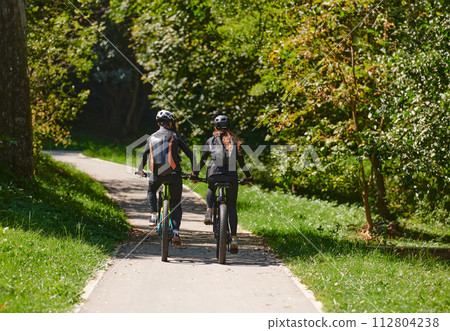 A blissful couple, adorned in professional cycling gear, enjoys a romantic bicycle ride through a park, surrounded by modern natural attractions, radiating love and happiness 112804238