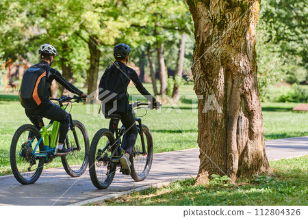 A blissful couple, adorned in professional cycling gear, enjoys a romantic bicycle ride through a park, surrounded by modern natural attractions, radiating love and happiness 112804326