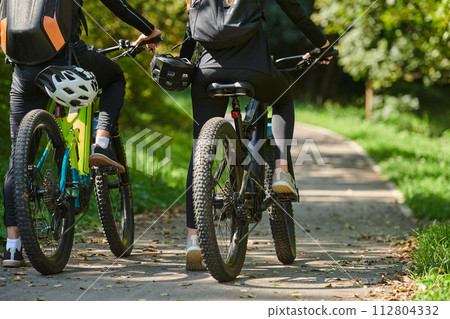 A blissful couple, adorned in professional cycling gear, enjoys a romantic bicycle ride through a park, surrounded by modern natural attractions, radiating love and happiness A blissful couple, adorned in professional cycling gear, enjoys a romantic bicycle ride through a park, surrounded by modern natural attractions, radiating love and happiness 112804332