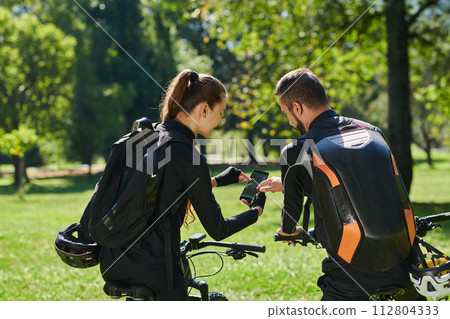 A sweet couple, equipped with bicycles and engrossed in coordinating their journey, checks their GPS mobile and watches while planning scenic routes in the park, seamlessly blending technology and 112804333
