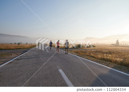 A group of friends, athletes, and joggers embrace the early morning hours as they run through the misty dawn, energized by the rising sun and surrounded by the tranquil beauty of nature A group of friends, athletes, and joggers embrace the early morning hours as they run through the misty dawn, energized by the rising sun and surrounded by the tranquil beauty of nature 112804334
