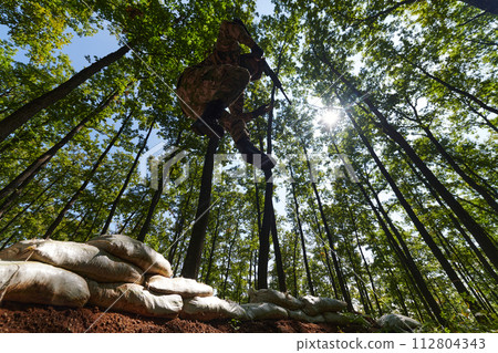 An elite soldier adeptly clears military barriers in the perilous wooded terrain, showcasing tactical skill and agility during specialized training An elite soldier adeptly clears military barriers in the perilous wooded terrain, showcasing tactical skill and agility during specialized training 112804343