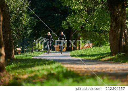 A blissful couple, adorned in professional cycling gear, enjoys a romantic bicycle ride through a park, surrounded by modern natural attractions, radiating love and happiness 112804357