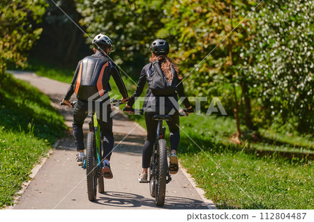 A blissful couple, adorned in professional cycling gear, enjoys a romantic bicycle ride through a park, surrounded by modern natural attractions, radiating love and happiness 112804487