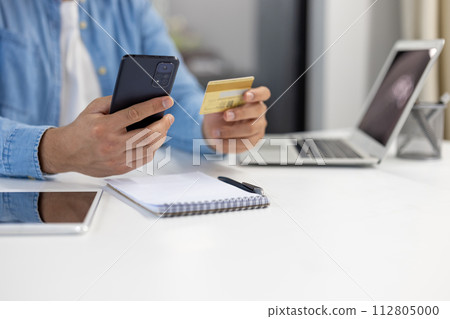 Close-up of a man's hands using a smartphone and credit card to make an online purchase with laptop and notebook in background. 112805000