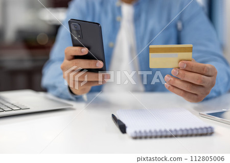 Close-up of a male's hands using a smartphone and credit card for an online transaction on a white desk with a notepad. 112805006