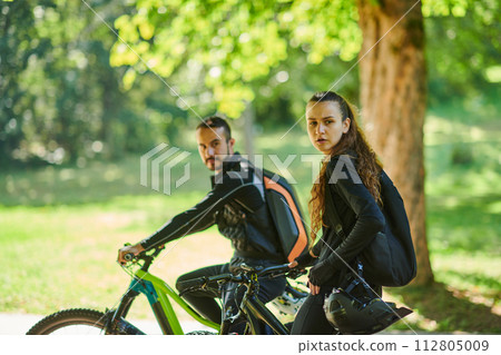 A blissful couple, adorned in professional cycling gear, enjoys a romantic bicycle ride through a park, surrounded by modern natural attractions, radiating love and happiness 112805009