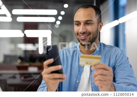 Confident young man holds a credit card and smartphone, shopping online with a smiling expression in a modern office setting. Confident young man holds a credit card and smartphone, shopping online with a smiling expression in a modern office setting. 112805014