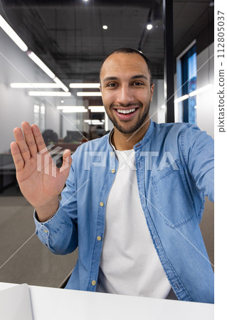 A young, cheerful businessman greets colleagues with a wave during a virtual meeting in a modern office setting. A young, cheerful businessman greets colleagues with a wave during a virtual meeting in a modern office setting. 112805037