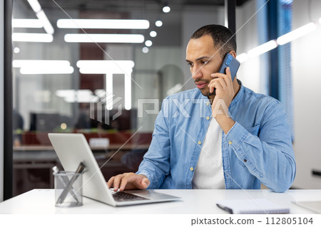 A focused professional man engaging in a phone conversation while working on a laptop in a modern office environment. A focused professional man engaging in a phone conversation while working on a laptop in a modern office environment. 112805104