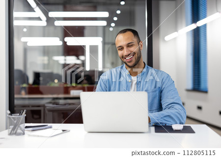 Happy young professional works on computer with a smile, in a bright contemporary workspace. Represents job satisfaction and a positive work environment. 112805115