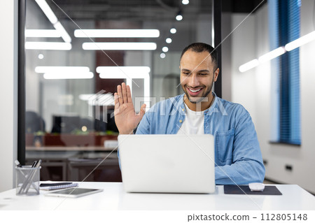 A cheerful businessman waves to the camera while engaging in a video conference in a modern office setting. A cheerful businessman waves to the camera while engaging in a video conference in a modern office setting. 112805148