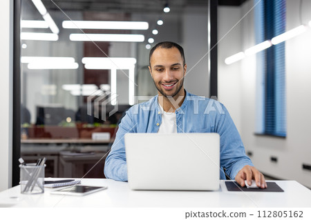 Smiling young professional man using laptop at his desk in a well-lit modern office, showing productivity and comfort. Smiling young professional man using laptop at his desk in a well-lit modern office, showing productivity and comfort. 112805162