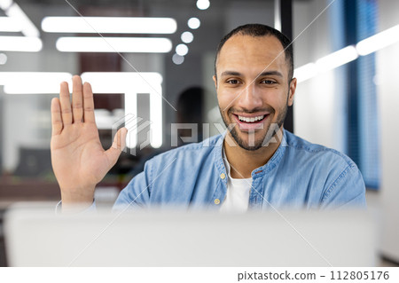 A cheerful young adult male waving at the camera during a casual video conference in a modern office setting. A cheerful young adult male waving at the camera during a casual video conference in a modern office setting. 112805176